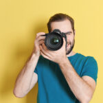 Photographer for Ocean County web design firm with camera up to eye standing in front of a yellow wall wearing a blue shirt